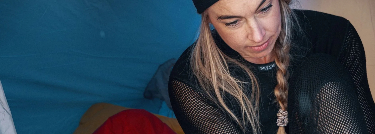 A woman with blonde hair in a braid, wearing a black beanie, black mesh long-sleeve base layer sitting inside a blue tent.