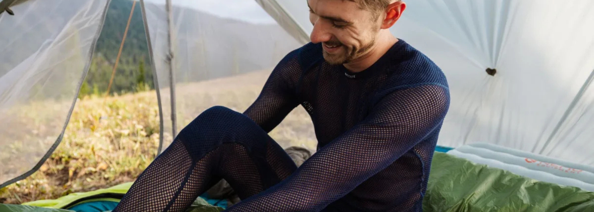 A man wearing a dark navy long-sleeve mesh base layer sits inside a tent in an outdoor mountain setting.