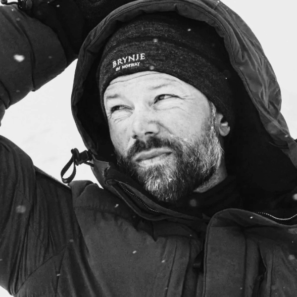 A black and white close-up of a man outdoors in the snow, wearing a dark hooded jacket and a knit beanie with the text 'BRYNJE OF NORWAY' visible on the front.